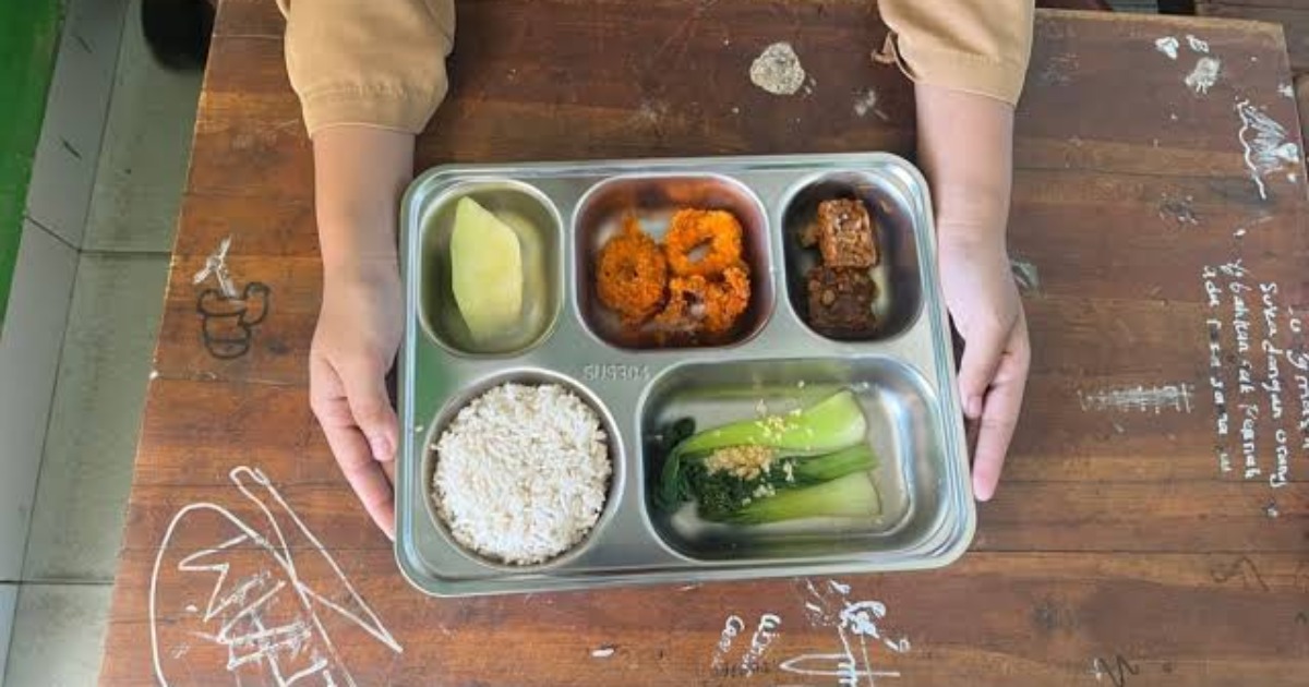 Siswa memegang nampan berisi menu Program Makan Bergizi Gratis (MBG) yang terdiri dari nasi, sayur, lauk, tempe, dan buah di ruang kelas.